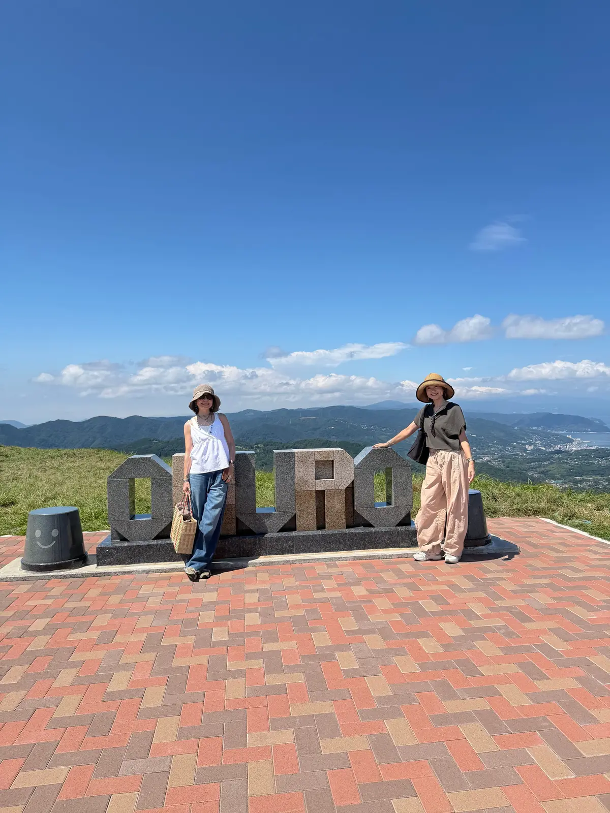山頂の展望台で、2人の女性が記念撮影をしている。背景には緑豊かな山々と遠くの街並み、青空が広がる。女性たちは麦わら帽子をかぶり、それぞれ異なる服装をしている。展望台には「OURO」と書かれた石碑が設置されている。