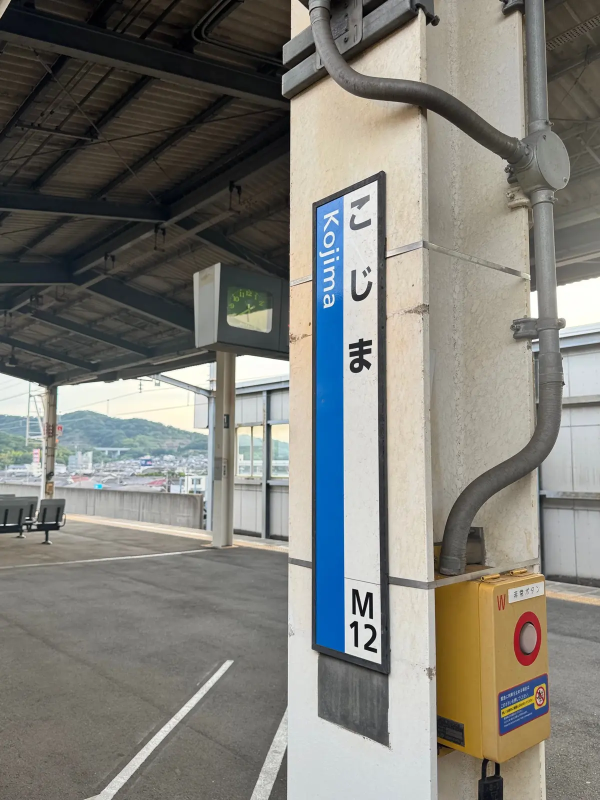 青い縦長の駅名標「こじま(Kojima)」、プラットフォームの風景。駅番号「M12」が記載。右には黄色い信号機と配管が設置。
