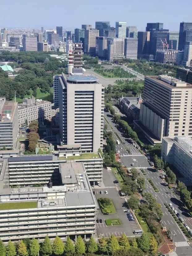 東京都庁舎を中心に、周辺のオフィスビルや公園、道路が広がる高層街並みの俯瞰写真。庁舎はベージュ色で、塔屋が特徴的。道路には車が走っており、緑豊かな公園が点在する。