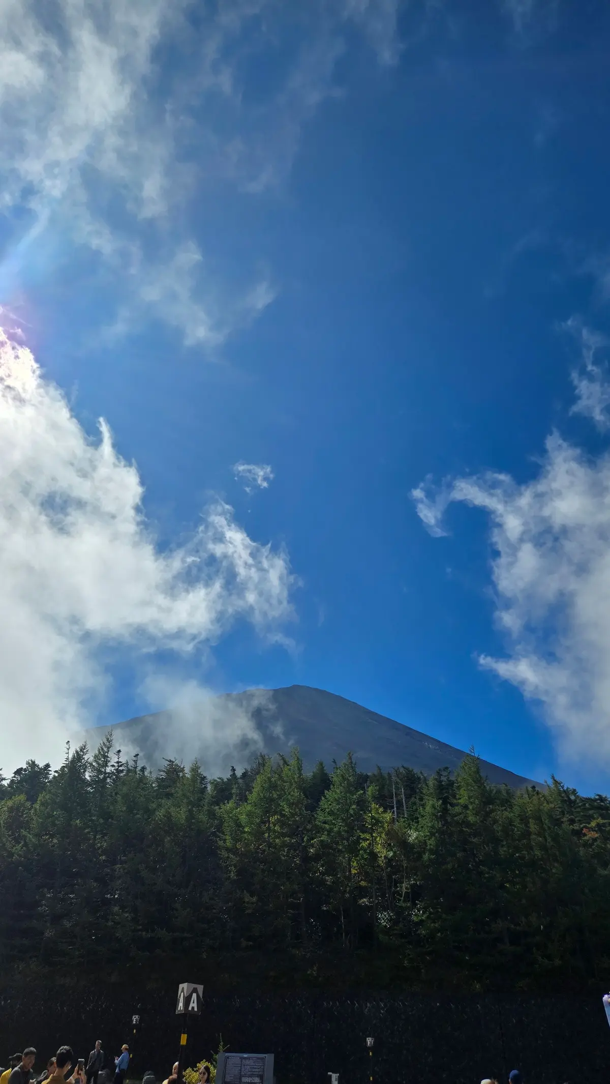 青空に白い雲、山頂付近は霧に包まれた富士山。手前には緑豊かな森林と、人々が写る。
