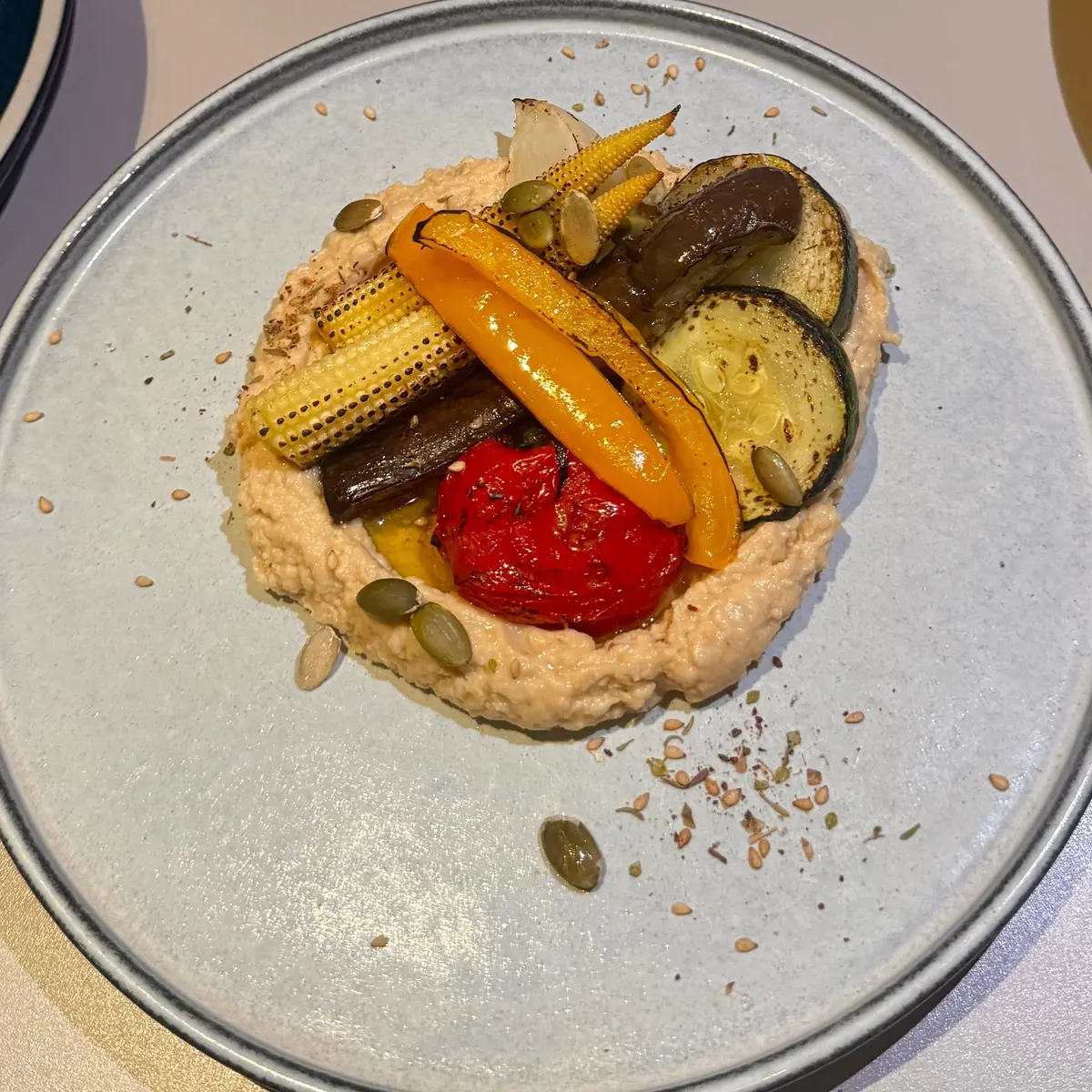 グレーの皿に盛り付けられた、ピンク色のホモゲニゼーションの上に、焼き野菜が彩り豊かに配置されている。赤、オレンジ、紫色の野菜が円形に並び、種子が散りばめられている。