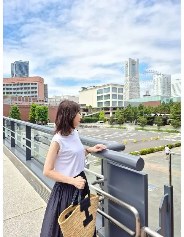 白いTシャツと黒いロングスカートを着た女性が、横浜の風景を眺めている。背景には、ランドマークタワーとコスモワールドの観覧車が見える。