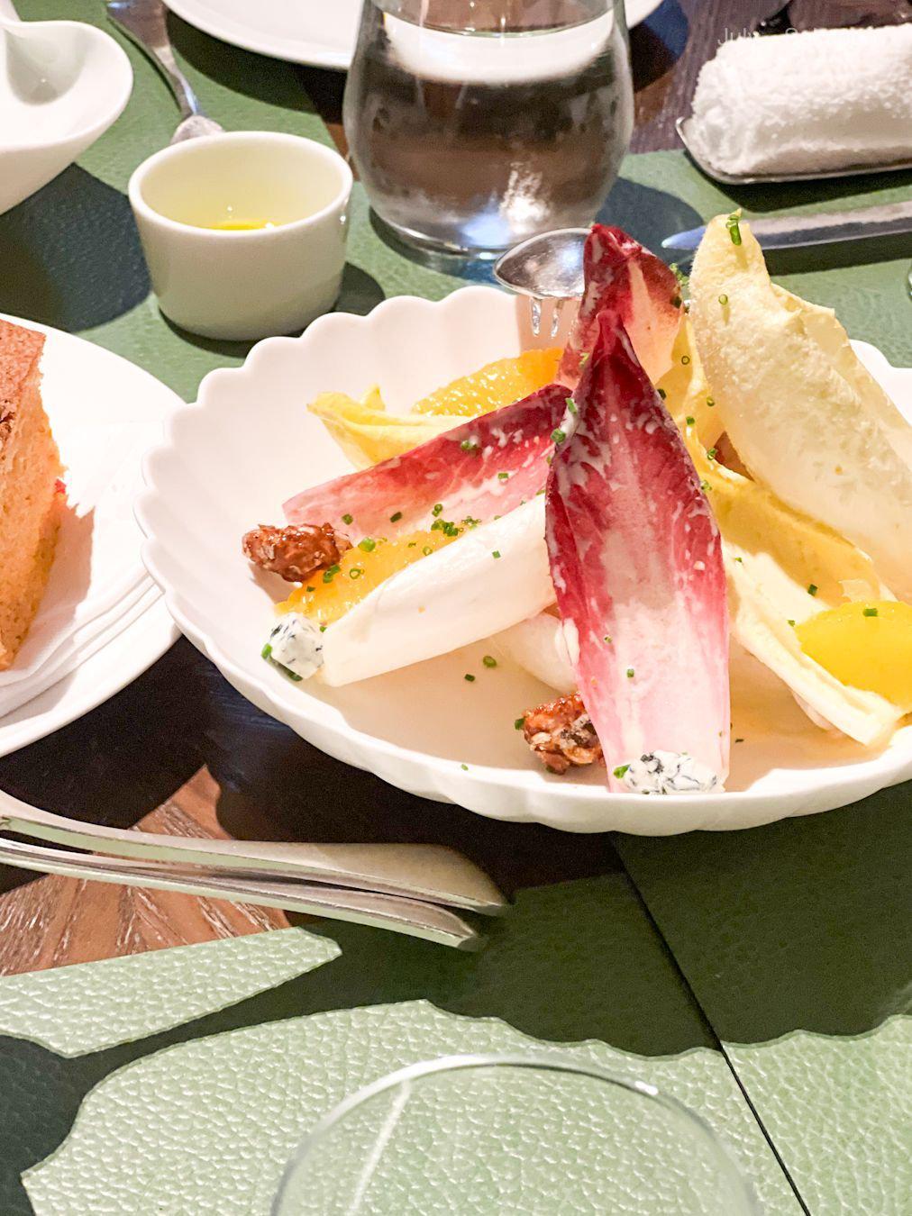 白磁の皿に盛り付けられたチーズの盛り合わせ。赤ジューシーな葉野菜、黄色い柑橘類、白いチーズが彩りを添えている。小皿には黄色のソース、グラスには水が置かれている。