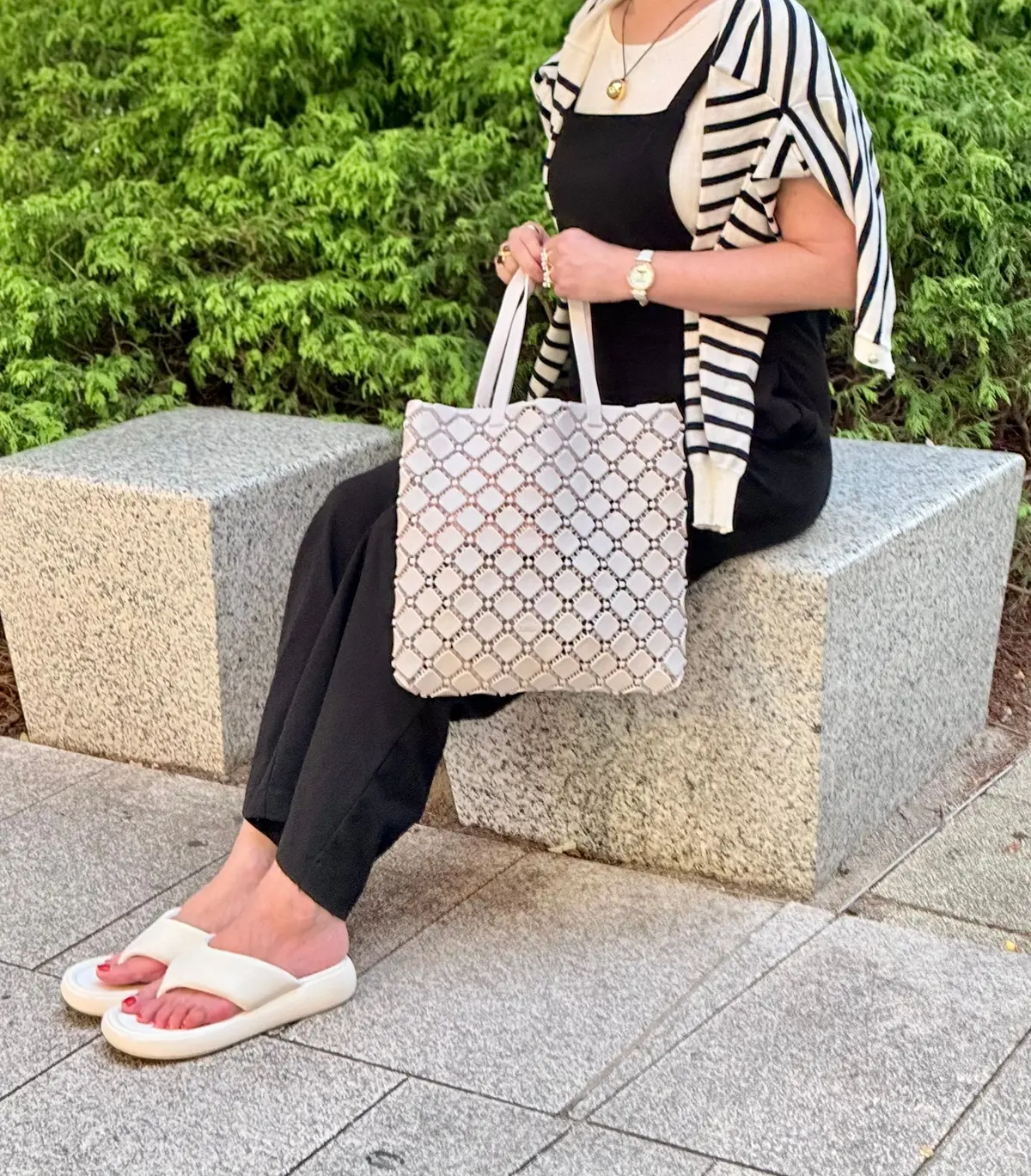 白いサンダルを履いた女性が、白いトートバッグを持って座っている全身写真。黒いパンツと黒いトップスに、白と黒のストライプのショールを羽織っている。背景には緑の植物が茂り、座っている場所はグレーの石造りのベンチ。