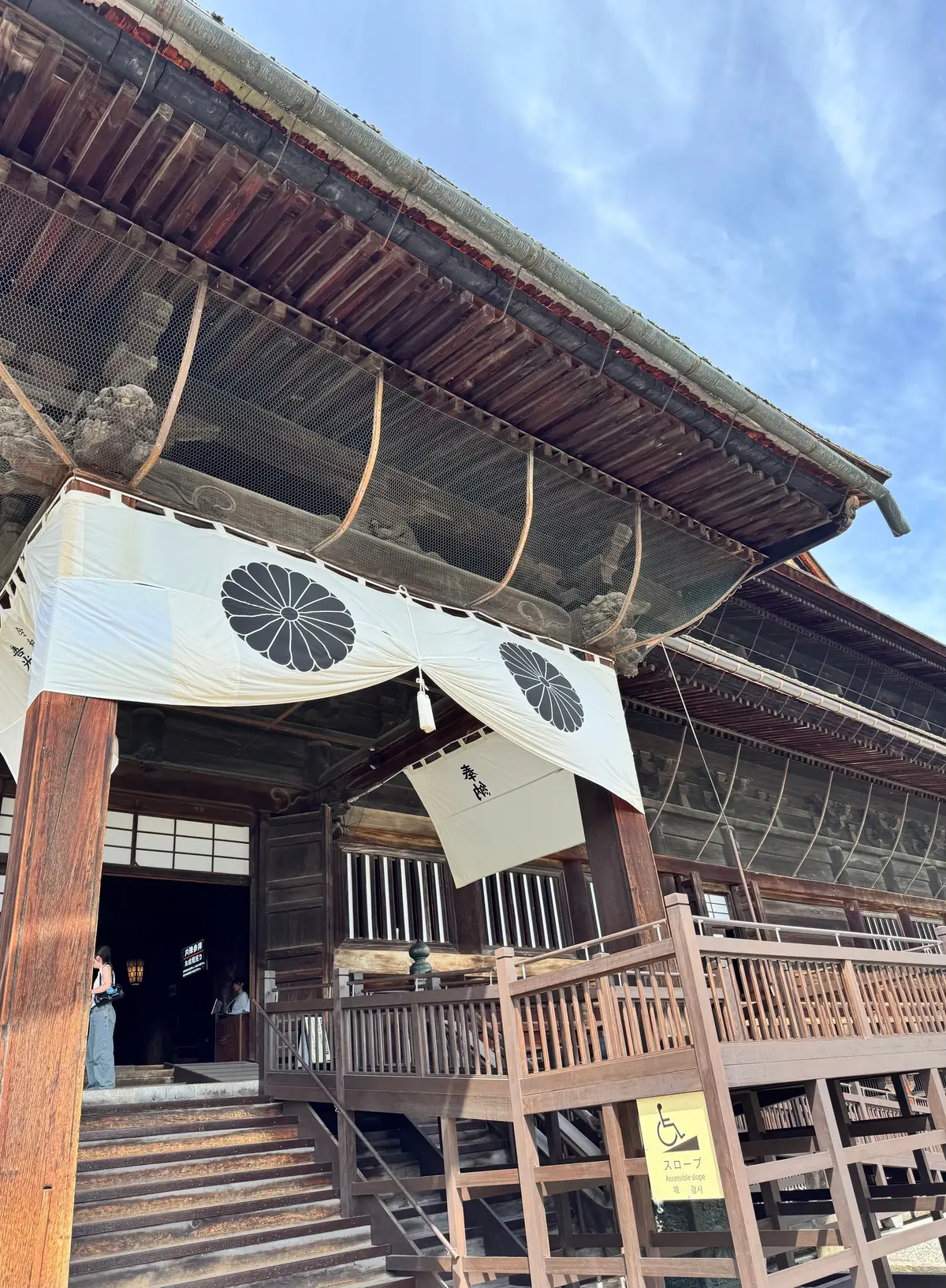 木造の伝統的な建造物の屋根と、白地に黒い模様が描かれた垂れ幕。手すりのある階段があり、バリアフリーマークの標識が設置されている。