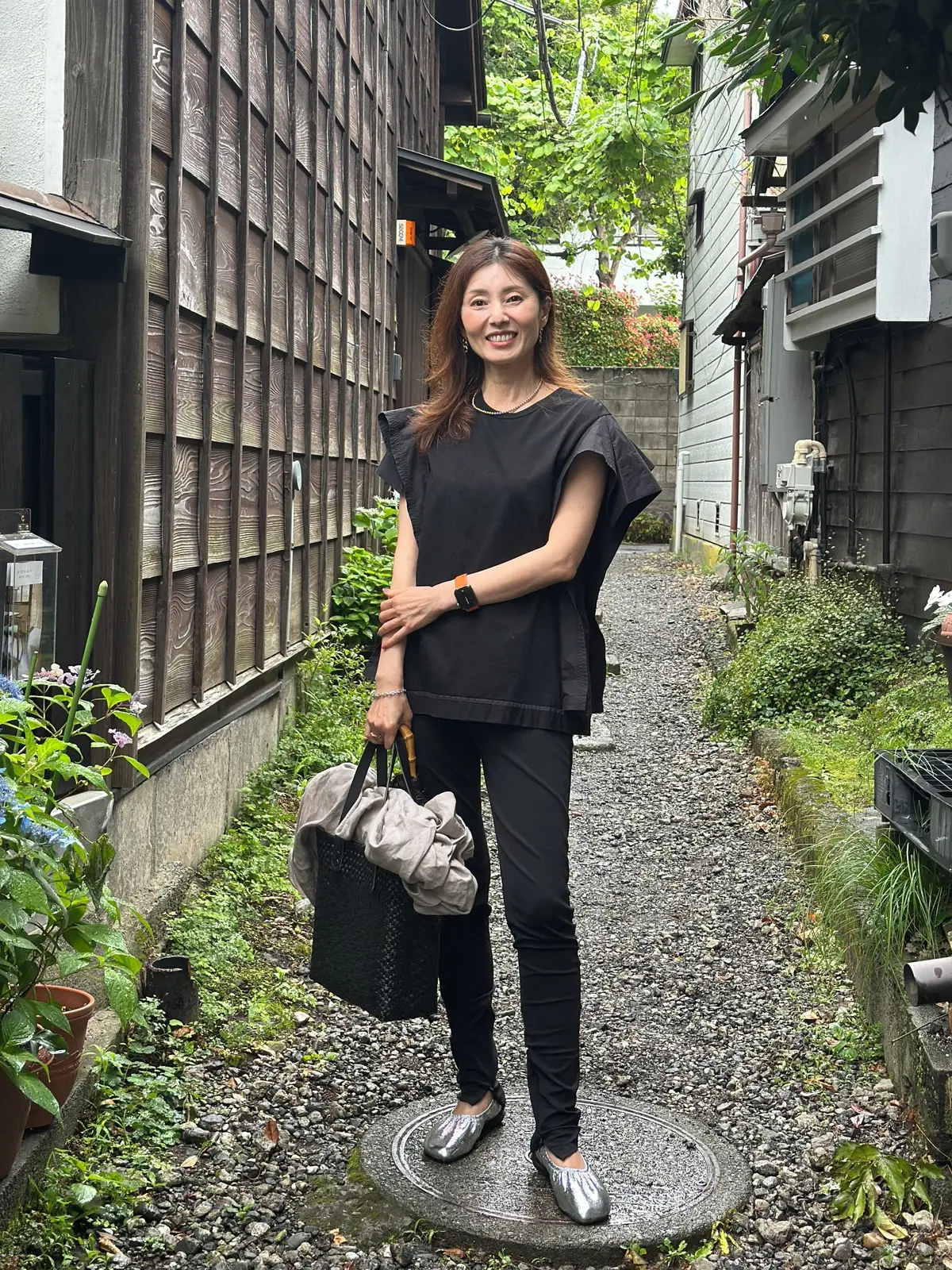 黒いトップスとパンツを着た女性が、石畳の路地に立っている。右手にベージュのバッグを持ち、銀色の靴を履いている。背景には木造の建物と緑の植物が見える。