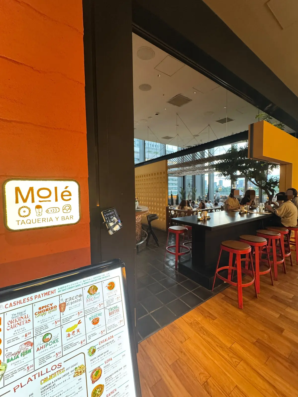 オレンジ色の壁に「Molé Taqueria y Bar」のロゴがあるレストランの店内。木製のテーブルと赤いスツールが並び、客が食事を楽しんでいる。窓から外の景色が見える。