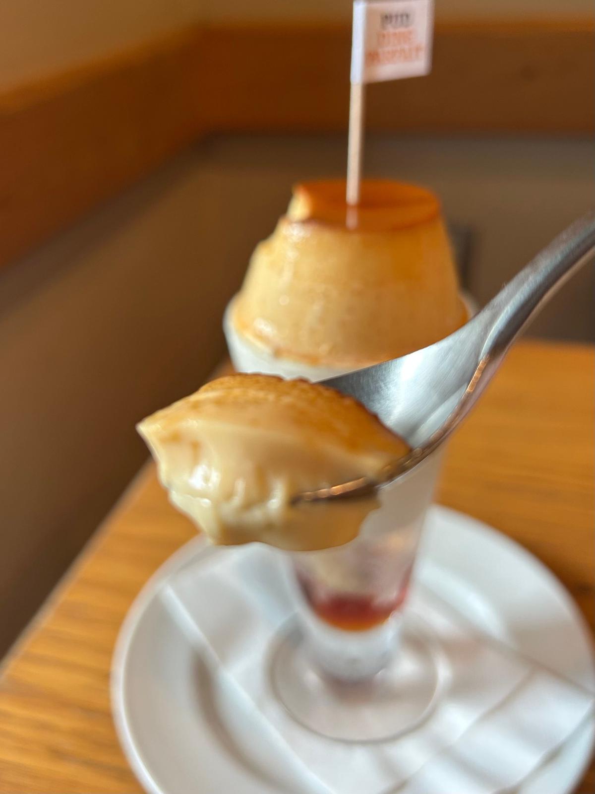 焦げ目がついたキャラメルソースをかけたクリームブリュレが、ガラスの器に入って白いお皿に置かれている。銀色のスプーンでブリュレをすくい上げている。