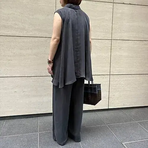 灰色のノースリーブシャツとワイドパンツのセットアップを着用した女性が、壁の前に立っている。女性は後ろ姿で、右手に茶色のバッグを持っている。