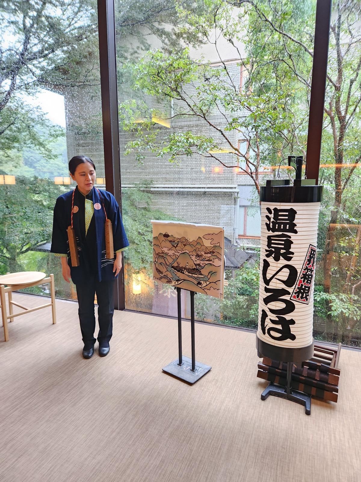 木目調の床に立つ女性が、黒と黄色の着物と灰色のズボンを着用。背景には窓から緑豊かな景色と建物が見える。右には白地に黒文字の提灯、手前には風景画が飾られている。