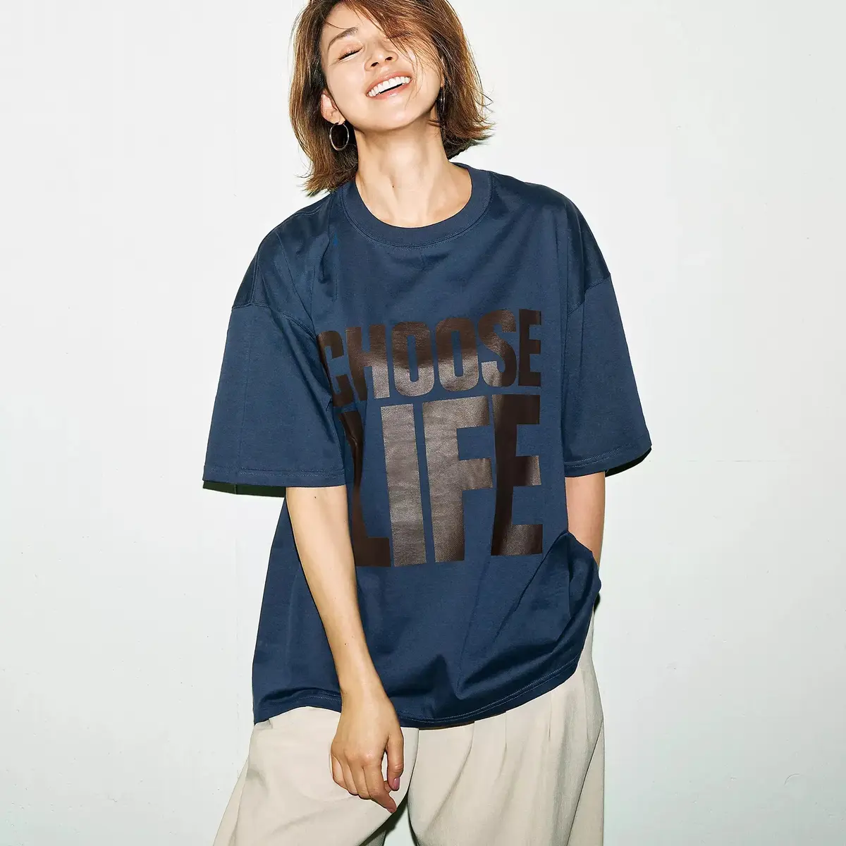 紺色のオーバーサイズTシャツを着た女性が、片手で裾を持ち、笑顔でこちらを見ている。Tシャツには茶色の文字で「CHOOSE LIFE」とプリントされている。ベージュ色のゆったりとしたパンツを着用。白い背景。