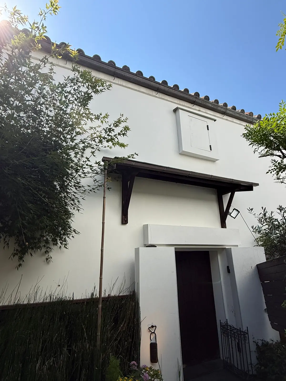 白壁の建物、黒い屋根付きの入り口と窓、緑の植物が茂る庭。青空が背景。