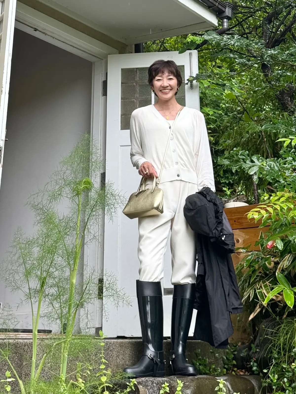白いワンピースと黒い長靴の女性が、屋外の階段に立ち、笑顔で金色のハンドバッグと黒い服を持っている。背景には緑豊かな植物と白いドアが見える。
