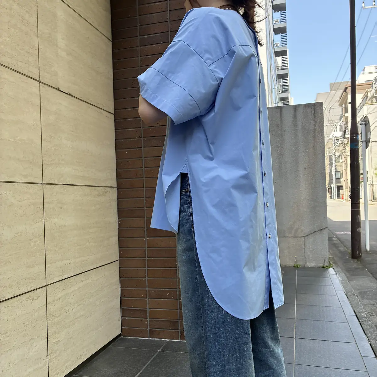軽い青のゆったりしたシャツを着た女性が、コンクリート壁の横で背面を向いて立っている。グレーのズボンを履き、建物と電柱が背景にある。