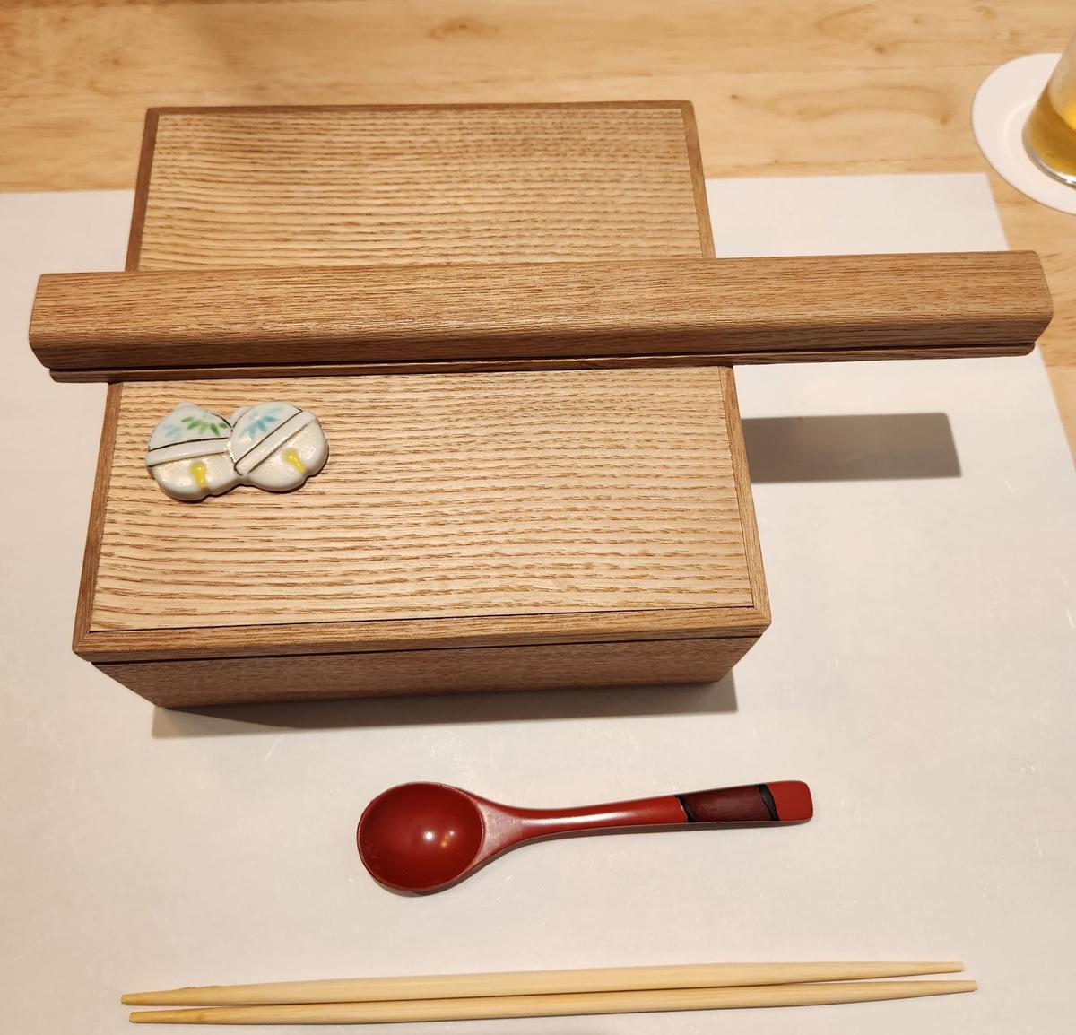 木製の弁当箱と、その上に置かれたお守り、赤い漆塗りのスプーン、木製の箸が置かれている様子。白いテーブルクロスの上に配置されている。
