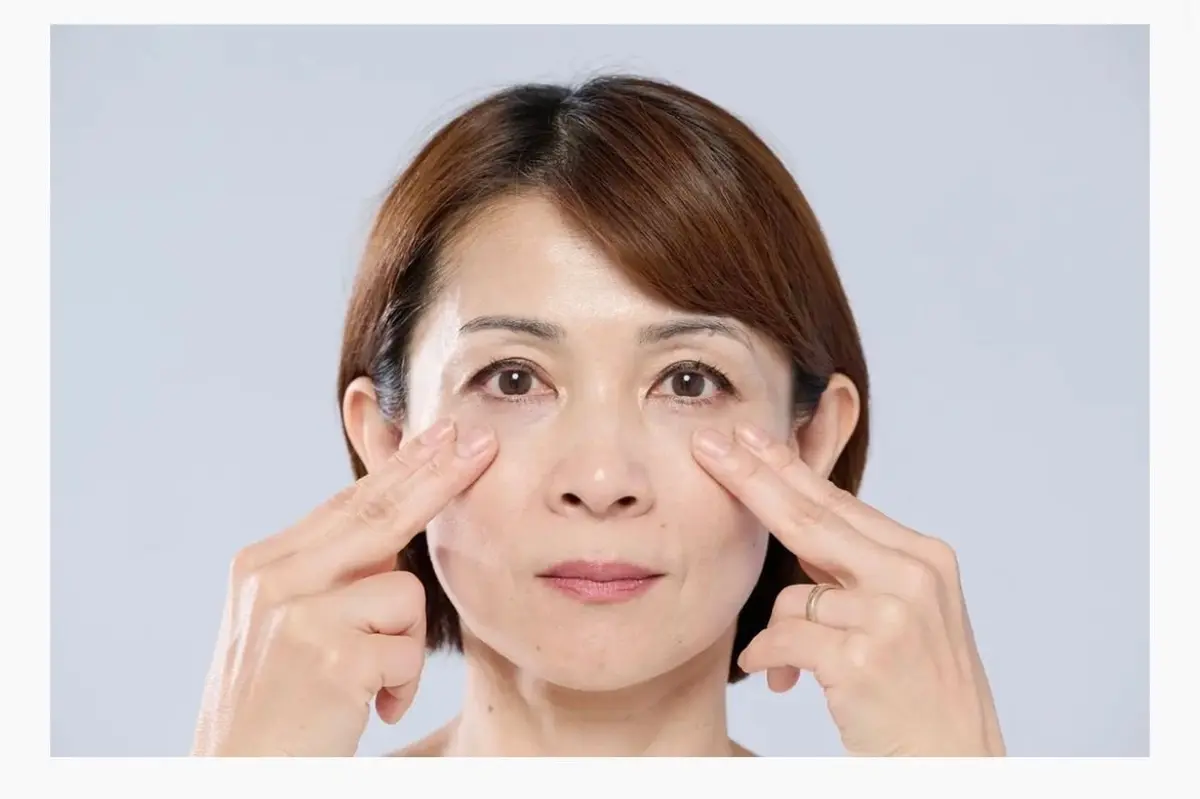 茶髪の女性が、両手で目の下を指で軽く押しているポートレート。背景は淡いグレー。