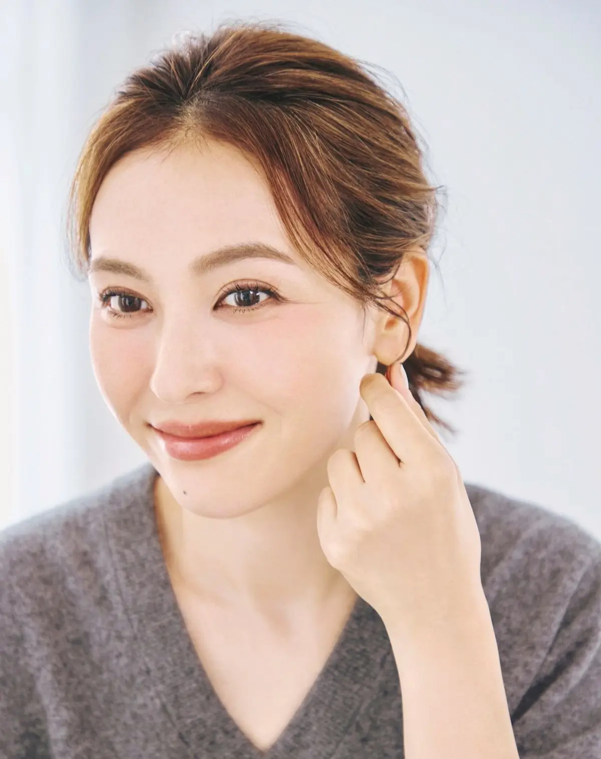 茶色の髪を後ろで一つにした女性が、微笑みながらこちらを見ているポートレート。グレーのセーターを着ており、肌は明るく、自然なメイクをしている。背景は白。