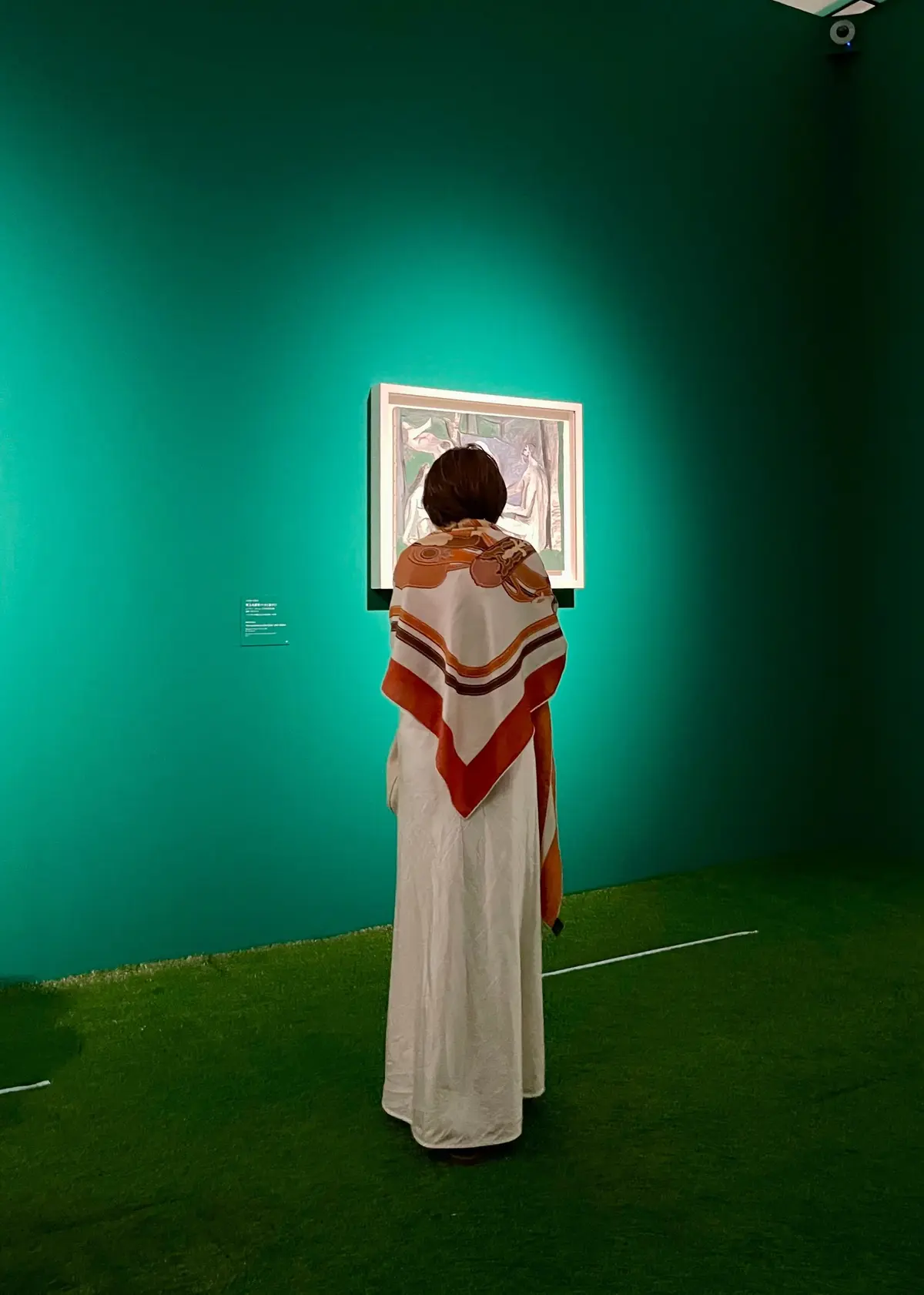 緑の壁に額装された絵画を、模様のあるスカーフを巻いた女性が後ろ姿で眺めている