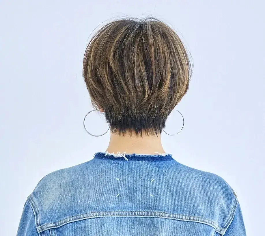 ショートカットの女性の後ろ姿で、茶色の髪が首元まで届き、大きな銀色のイヤリングを着用。デニムジャケットを着ている。