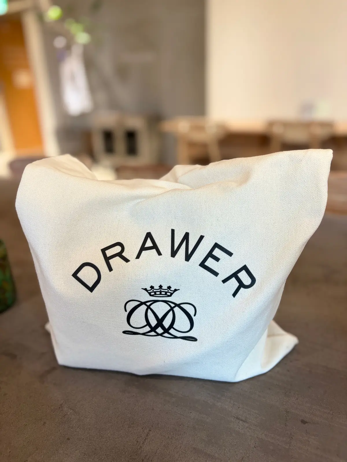 白いキャンバストートバッグ。中央に黒色の「DRAWER」の文字が弧を描くように配置され、その下に王冠と絡み合ったリボンのロゴマークがプリントされている。