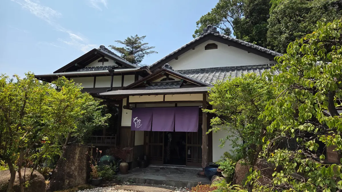 木々に囲まれた伝統的な日本の家屋。白い壁に瓦屋根、軒下には紫色ののれんがかかっている。入り口には木製の柱と格子。庭には石や植物が配置されている。