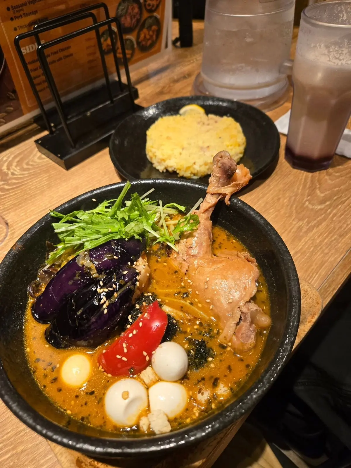 黒い丼に入ったラーメン。鶏肉、半熟卵、ネギ、赤と紫の野菜が盛り付けられている。右隣にはご飯、奥には飲み物が置かれている。