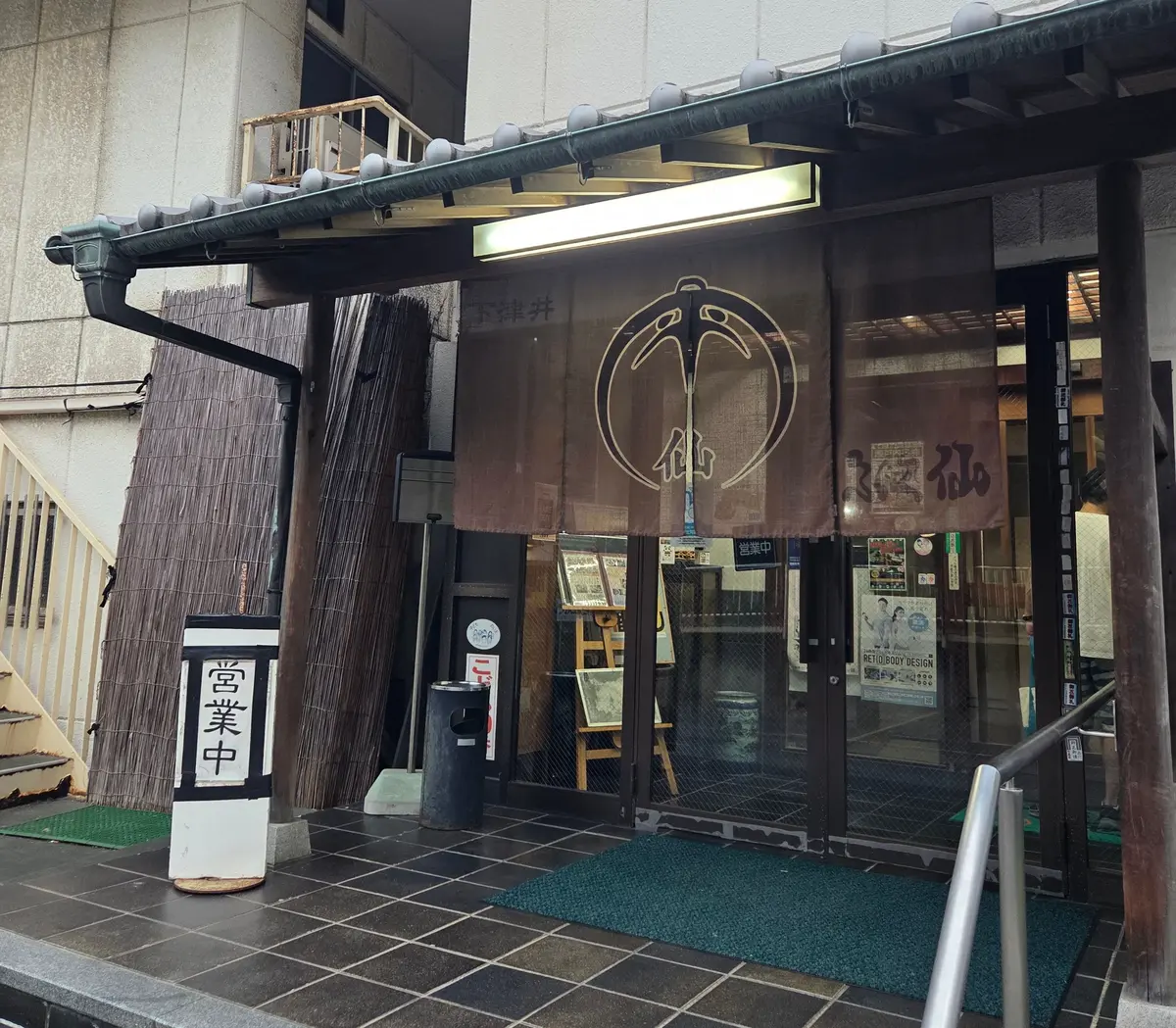 建物の入り口に設置された、竹や笠を模した装飾が施されたガラス張りの入り口。中央には鳥をかたどったロゴマークがある。白い手すりと、黒い柱が設置されている。