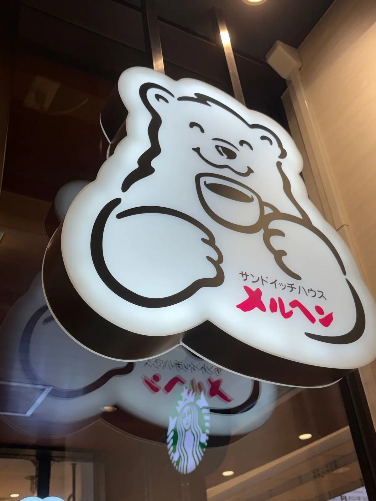白いクマのイラストが描かれた光るサイン。クマは片手を上げており、口にはコーヒーカップを持っている。サインの下には日本語の文字が書かれている。背景にはスターバックスの店舗が見える。