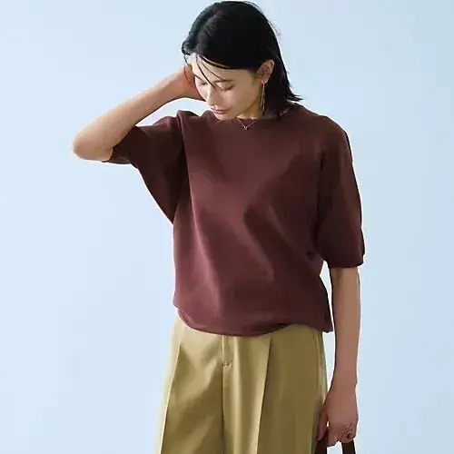 茶色のトップスにベージュのパンツを着用した女性が立っている写真。女性は右手を後頭部に当て、正面を向いている。背景は淡いブルー。