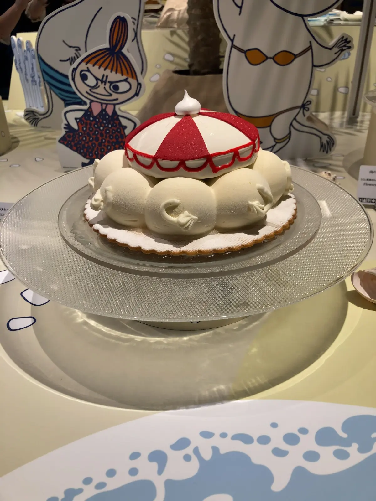 ムーミンキャラクターが描かれた紙製の立体ディスプレイと、赤と白の傘が乗ったムースケーキが並ぶテーブル