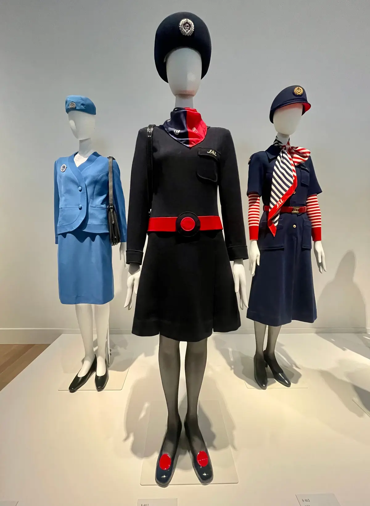 3体のマネキンが、日本航空（JAL）の歴代制服を着用して並んでいる。左から青色のスーツ、中央は黒と赤のワンピース、右は青と黄色のマリン風制服。各マネキンは帽子をかぶり、異なるデザインのネクタイやスカーフを着用している。背景は白。