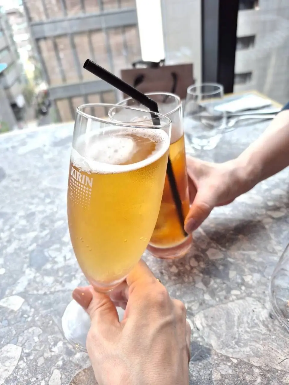 グレーのテーブルに置かれた、キリンのビールグラス2つ。泡が立ち、黒いストローが挿入されている。背景には窓と、一部のテーブルセッティングが見える。