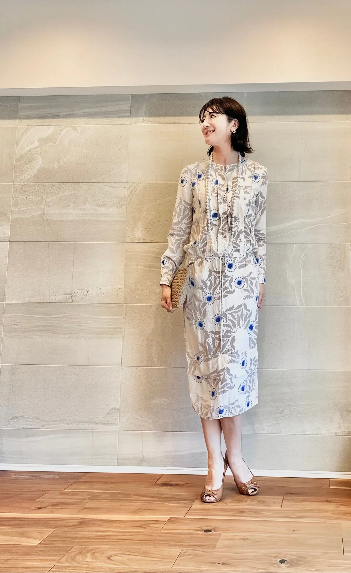 白い地に青い花柄のシャツワンピースを着た女性が、壁の前で微笑んでいる。茶色のハイヒールを履き、淡い色のネックレスをしている。