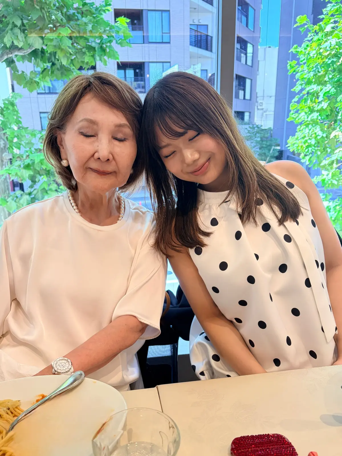 笑顔の女性2人が、レストランで食事中に寄り添って写るポートレート。左は上品な白のブラウスを着た年配女性、右は白いドット柄のワンピースを着た若い女性。背景には高層ビルと緑の木が見える。