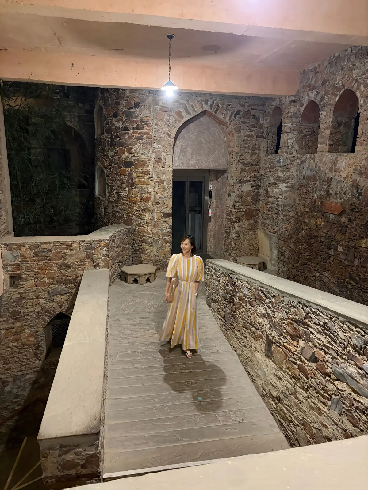 石造りのアーチが並ぶ古都の建造物の中を、黄色と白のストライプのワンピースを着た女性が歩いている。木製の通路が設けられ、奥には黒い扉が見える。