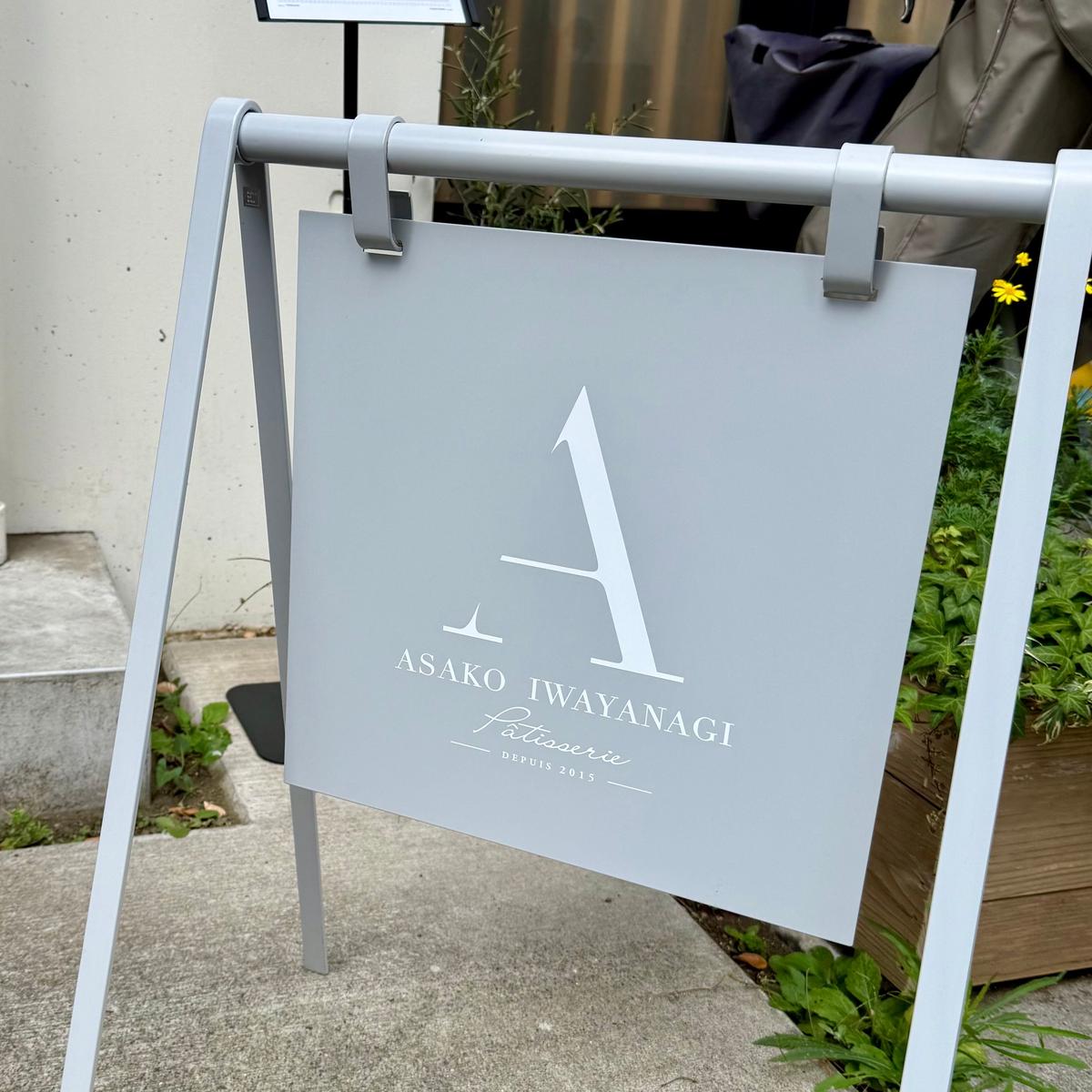 グレーのA型看板に、白抜きの数字「1」と「ASAKO IWAYANAGI Patisserie」の文字がデザインされている。看板の下部には「DEPUIS 2015」と記載されている。背景には緑の植物が見える。