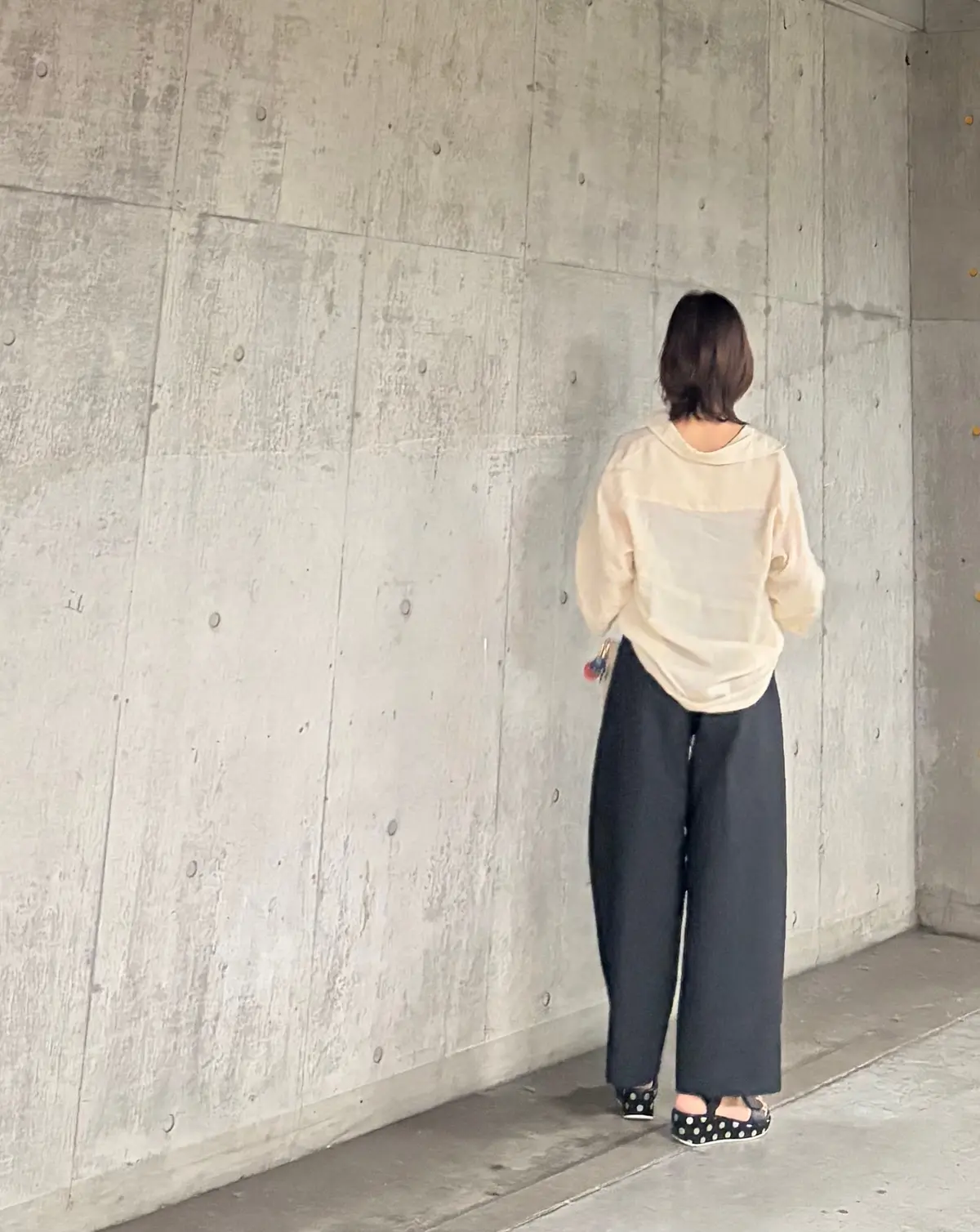 コンクリート壁に背を向けた女性、ベージュの長袖トップスと黒いワイドパンツ、ドット模様のサンダルを履く
