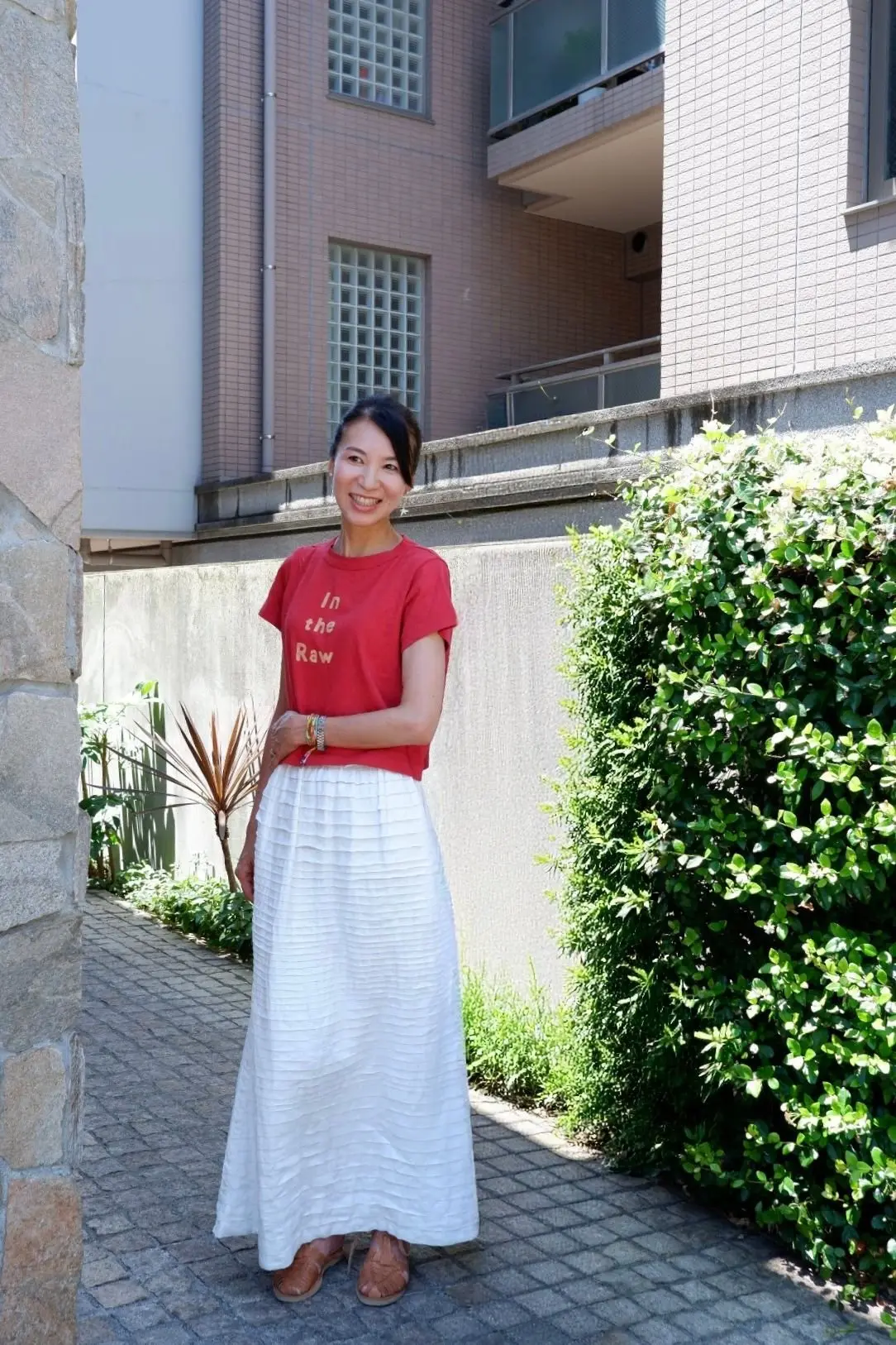 赤いTシャツと白いロングスカートを着た女性が、石積みの壁と緑の植栽を背景に立っている。笑顔でこちらを見ている。