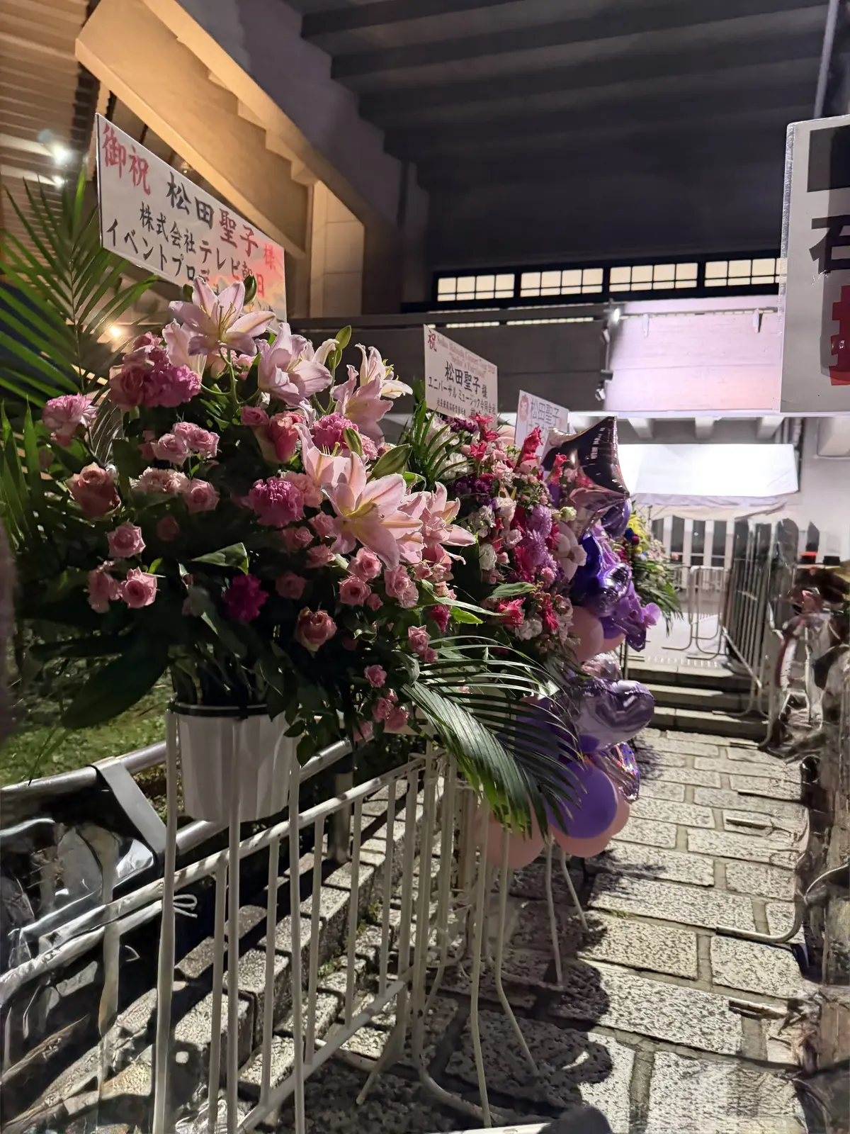 夜の石造りの建物前に、ピンクや白の生花、風船、メッセージカードが飾られている様子。手すりの向こうに、人影も見られる。