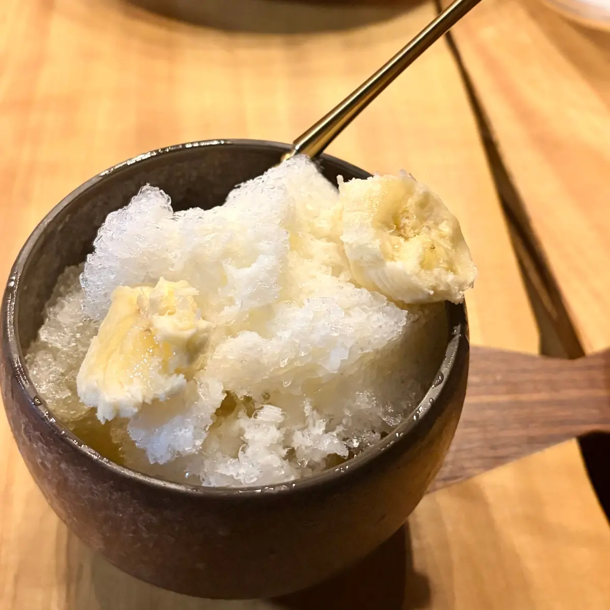 茶色の陶器の器に入ったかき氷に、バナナが添えられている。かき氷は細かく、バナナはスライスされている。木製のテーブルの上に置かれている。