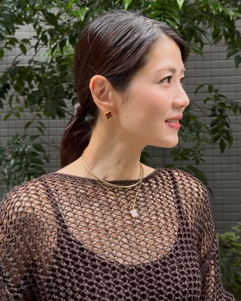 茶色の網目状のトップスを着た女性が横を向いている。黒髪を後ろでまとめ、金色のイヤリングとネックレスを身に着けている。背景には緑の葉とグレーの壁がある。