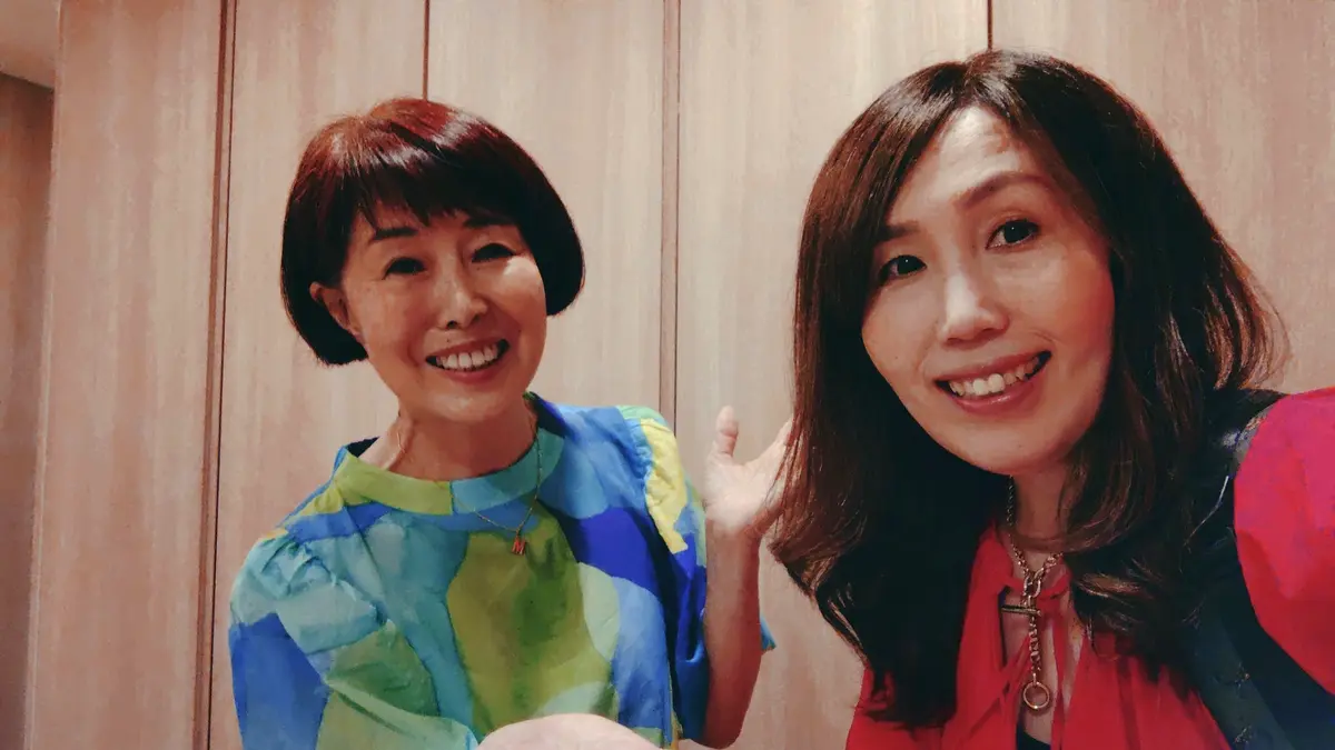 女性二人が並んで微笑んでいる。左の女性はショートヘアで青緑の模様のある服を着て、右の女性はロングヘアで赤の服を着ている。背景は木製パネルの壁。