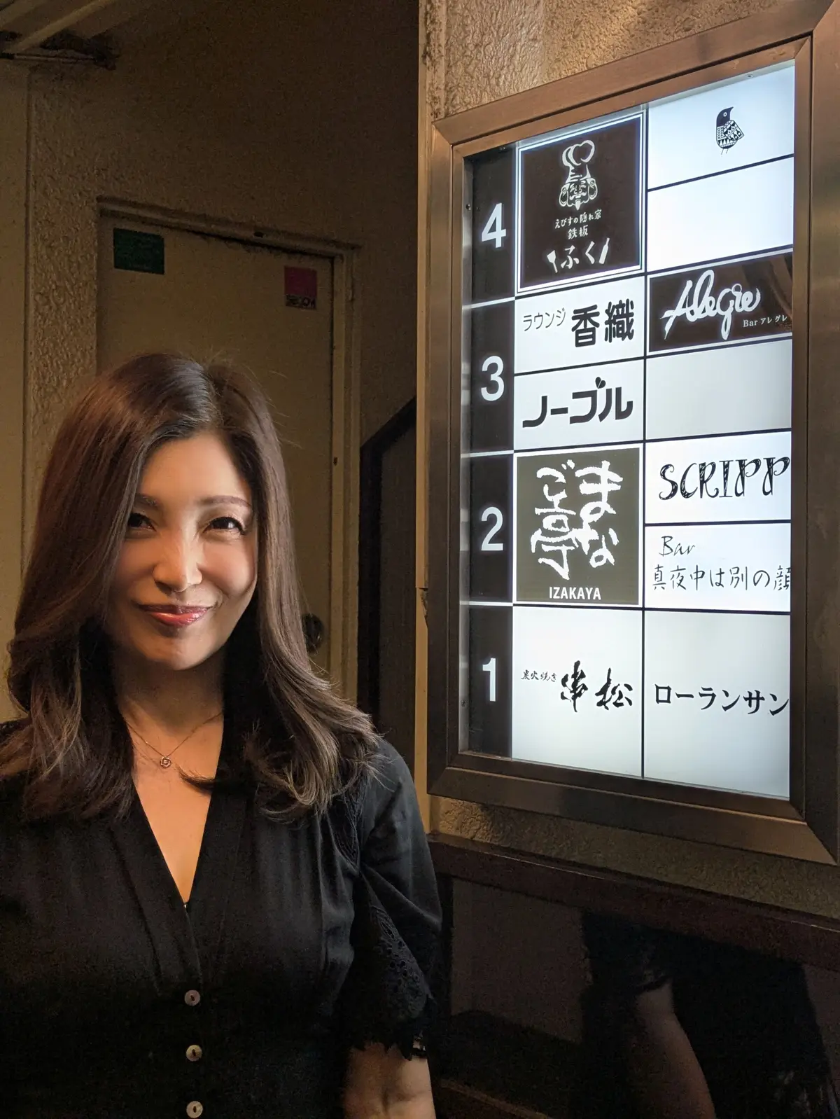 女性が黒い服を着て、4つのフロアを表す案内板の前で微笑んでいる。案内板には日本語と英語で「ローランサン」「イザカヤ」「ル・ベル」「クラシック」などの店名が記載されている。