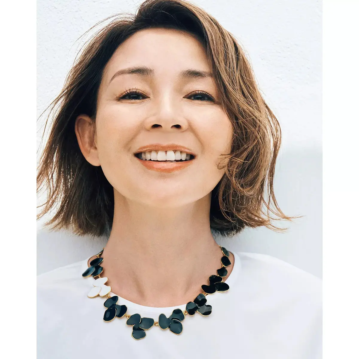黒のレースシャツと白いパンツを着た女性が、笑顔でドアの前で立っています。右手にはストローハンドバッグを持っています。
