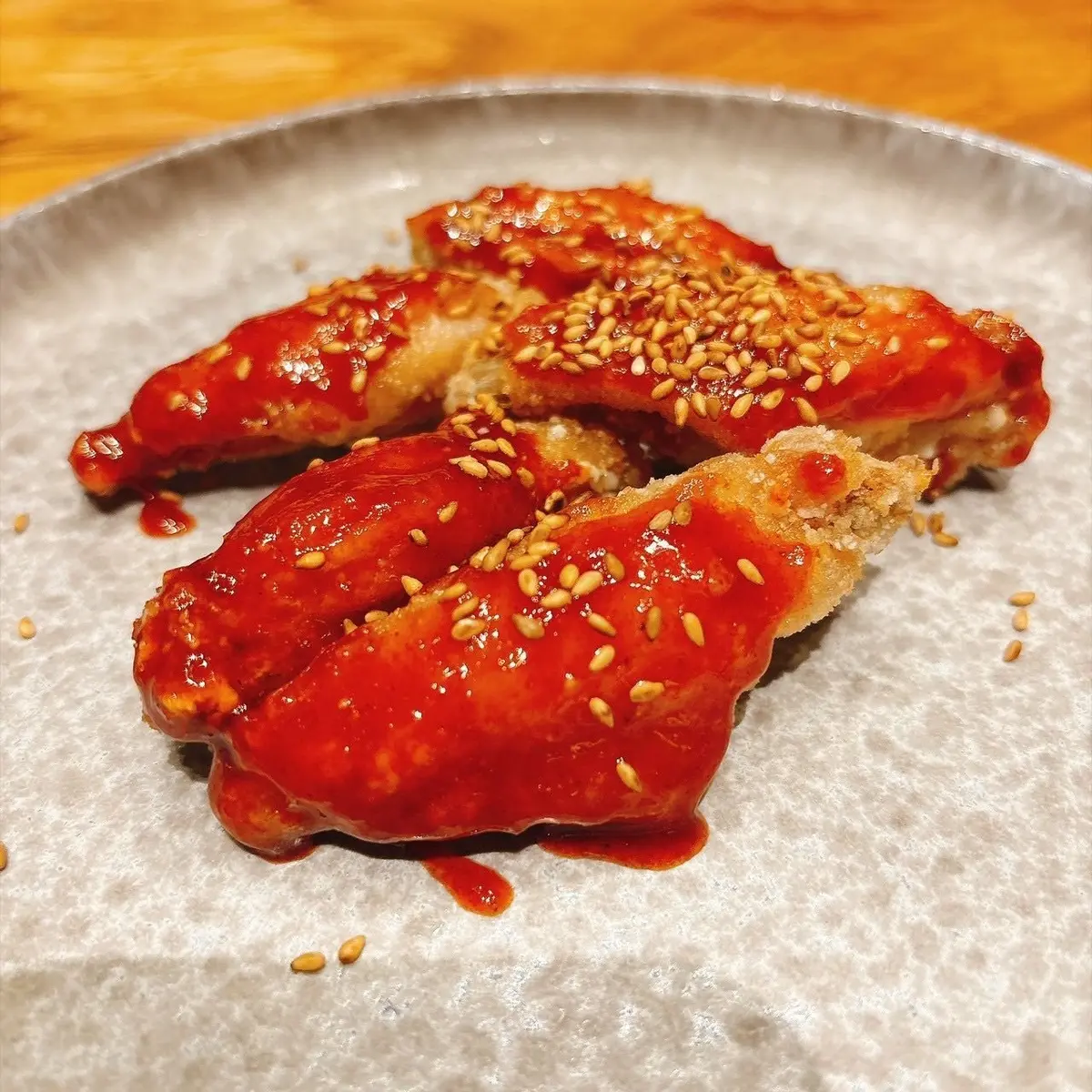 赤い甘辛ソースと白ゴマをかけた、揚げた鶏肉の切れ端が3つ、グレーの皿に盛り付けられている。