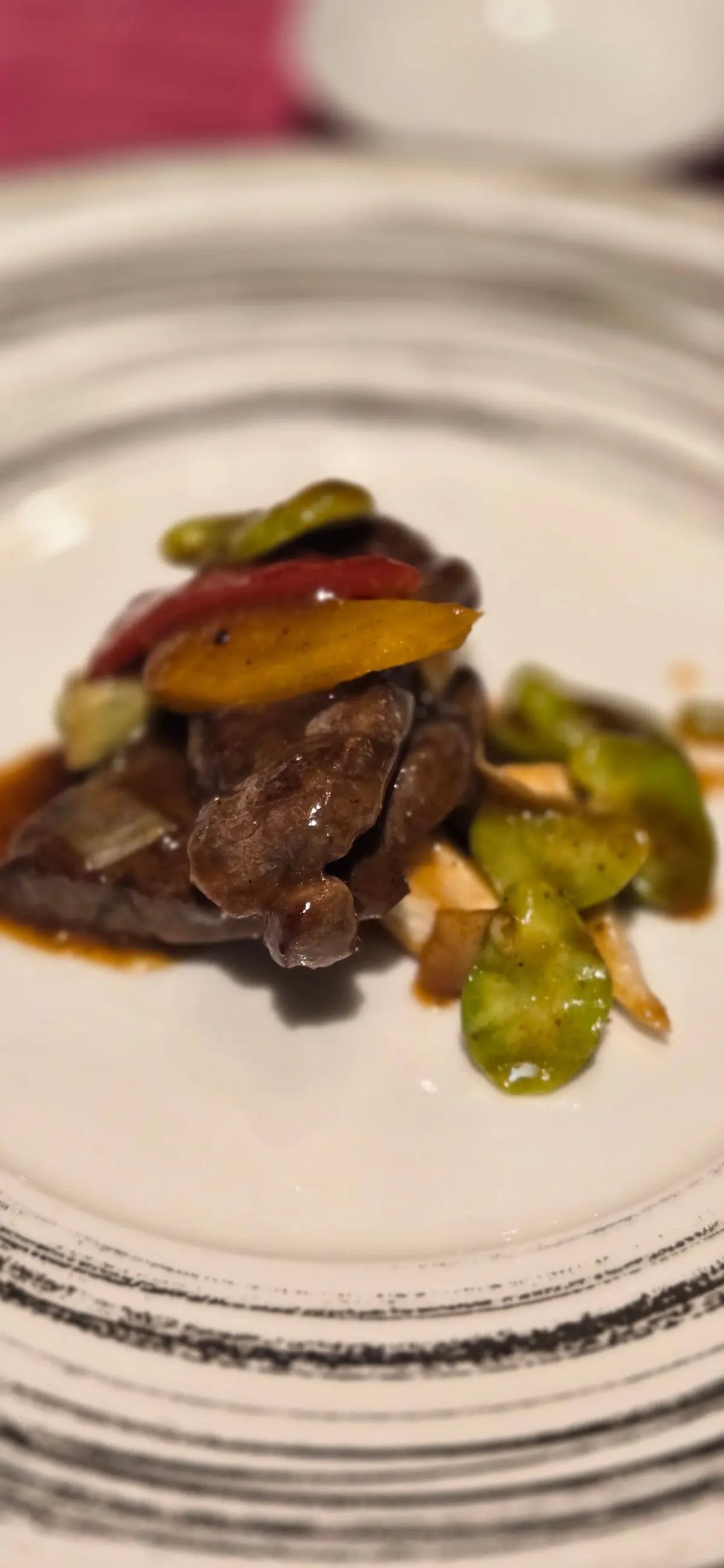 白磁の皿に盛り付けられた、牛肉と野菜の炒め物。赤と黄色のパプリカ、緑色のピーマン、玉ねぎが添えられ、濃い色のソースが注がれている。