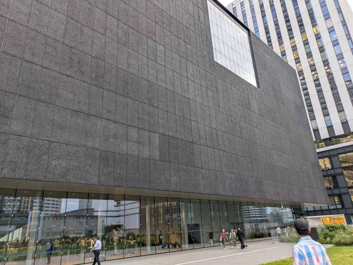 現代的なデザインの灰色の建物と、隣接する白と黒のストライプのオフィスビル。建物の正面にはガラス張りの入り口があり、人々が歩いている。