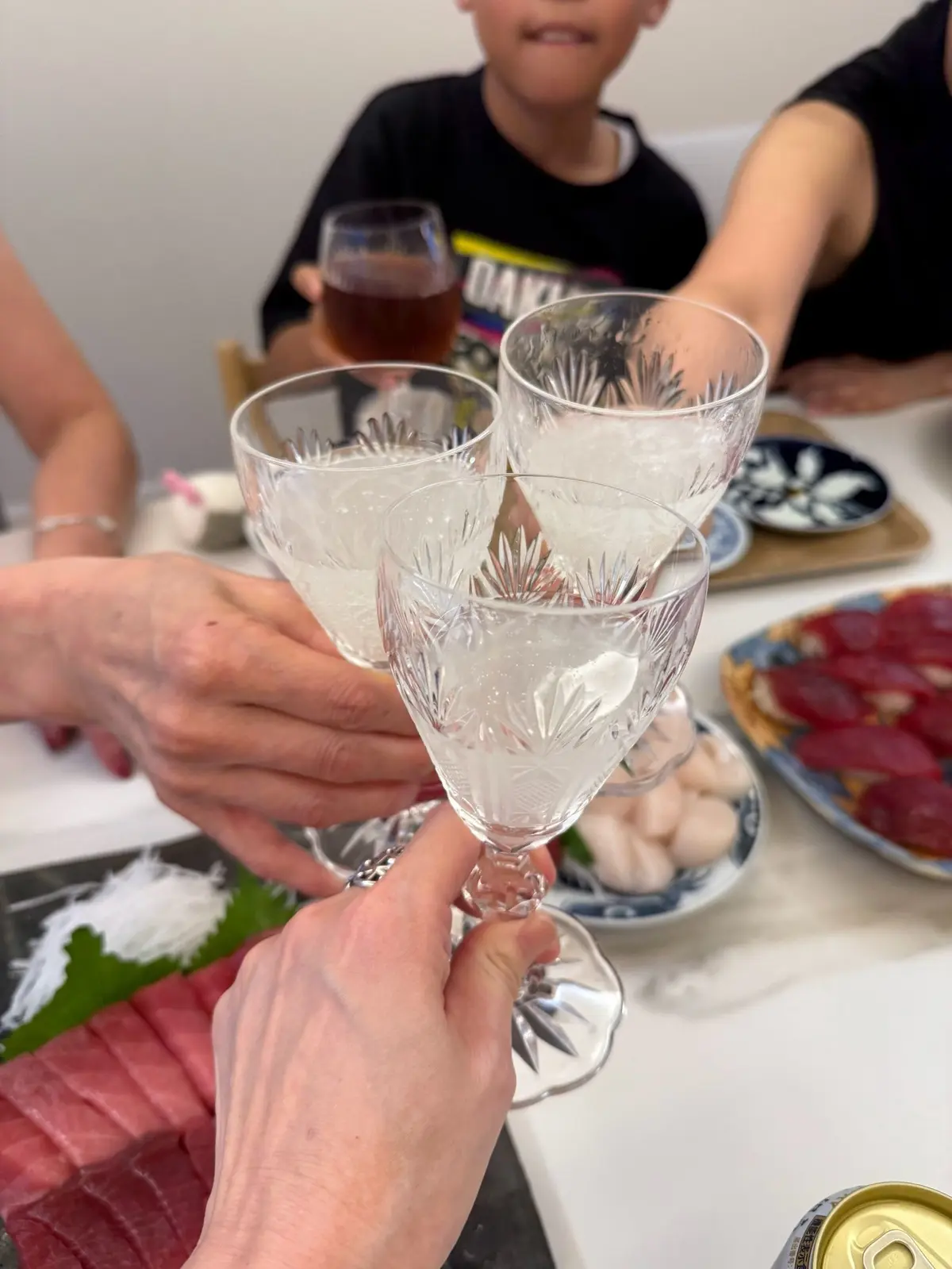 透き通ったグラス4つが重なり合い、乾杯の様子。背景には、ビール、料理、子供の姿が見える。テーブルには、緑色の野菜や赤い肉料理が並んでいる。