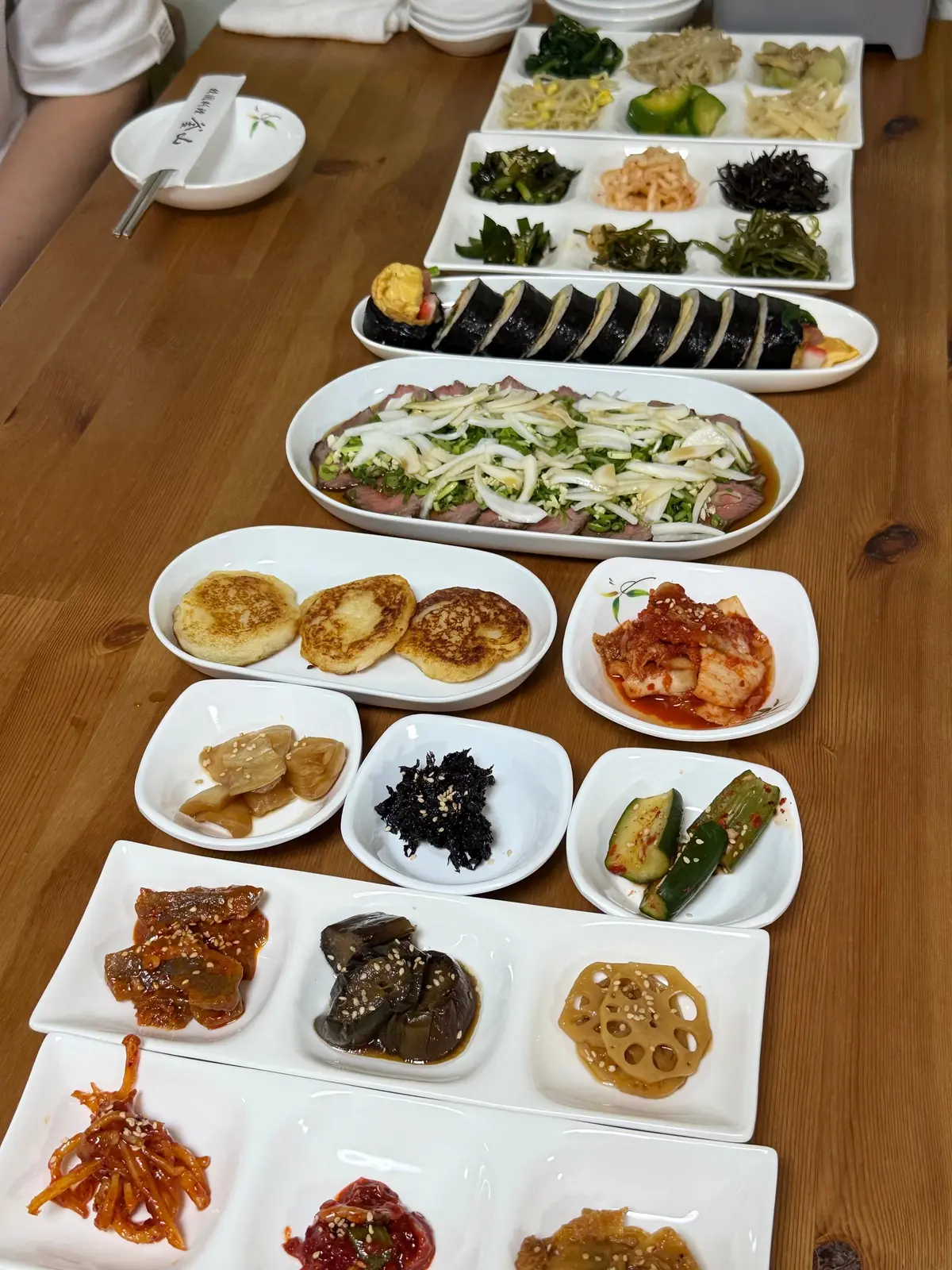 韓国料理 釜山 予約が取れないお店　尼崎グルメ　おすすめ　パンチャン　パンチャン　木のテーブルにさまざまな韓国料理が並ぶ。肉の盛り合わせ、焼いたパン、キムチ、海藻、野菜の漬物、卵焼きなど多彩な料理が白い皿に盛り付けられている。