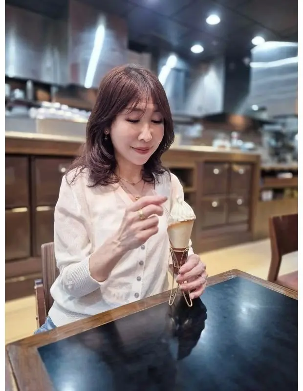 カフェのような場所で、女性がソフトクリームのコーンを持っている。茶色の髪、白いトップスを着用し、少し微笑んでいる。背景には木製の収納棚が見える。
